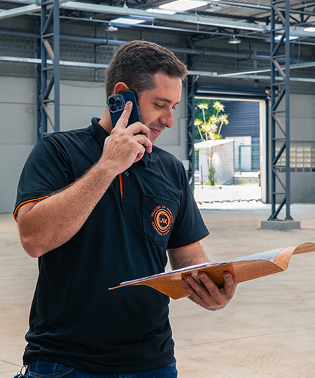 Representante técnico em visita consultando documentação e falando ao celular em galpão industrial — coordenação de projetos e atendimento técnico