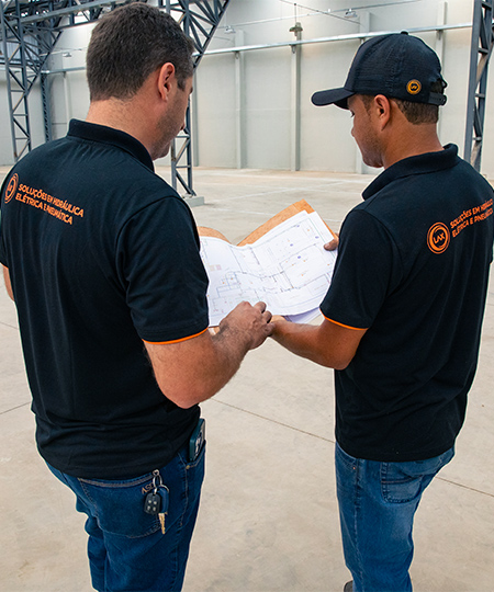 Dois técnicos da LAX em uniforme consultando plantas técnicas em prancheta dentro de galpão